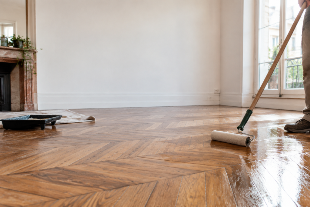 Une personne applique du vernis sur un parquet en chevrons ancien à l'aide d'un rouleau à long manche.